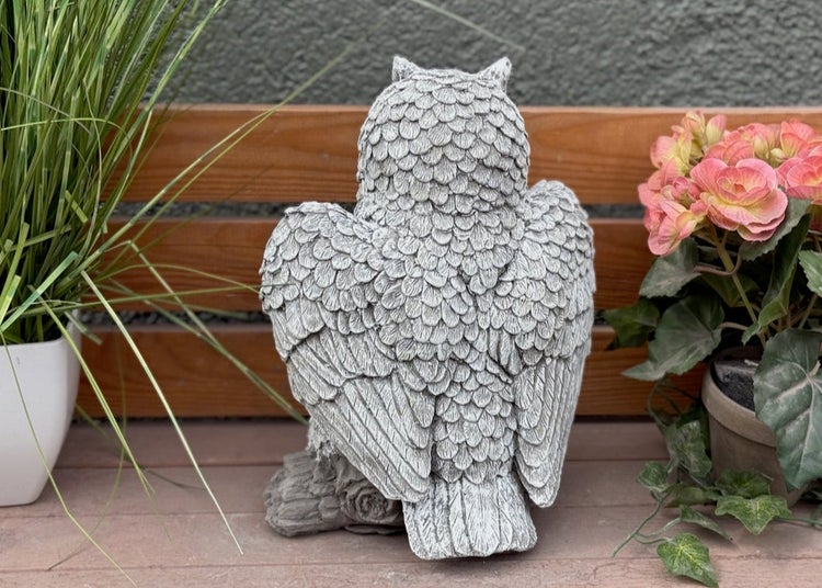 Steinfigur Eule in detaillierter Gefiederoptik als Gartendekoration auf einem Holzuntergrund zwischen Topfpflanzen.