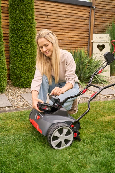 Eine Frau setzt einen Akku in einen grau-roten Akku-Vertikutierer auf einem Rasen in einem Garten mit Holzzaun und Büschen ein.