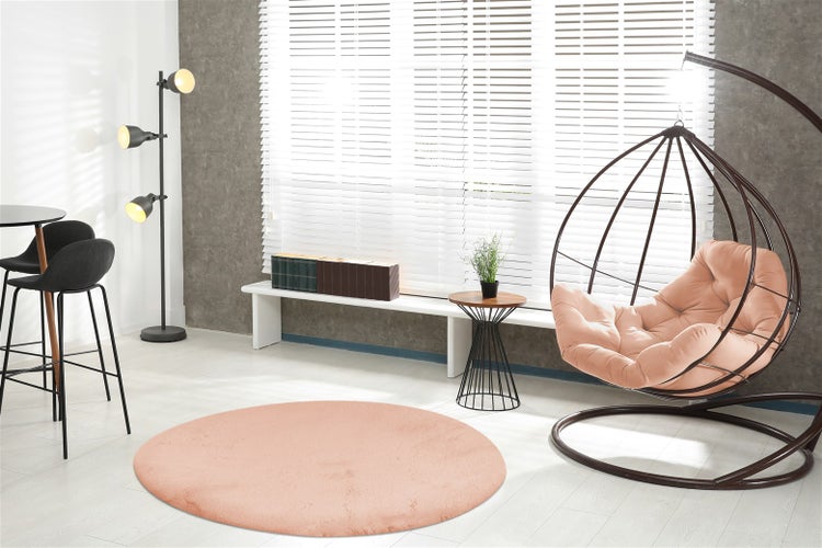 Modernes, helles Wohnzimmer mit einem runden, pfirsichfarbenen Teppich, einem Hängesessel, weißen Jalousien und einer Stehleuchte.