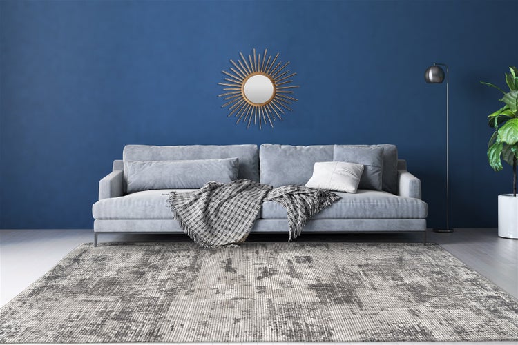 Wohnbereich mit abstraktem Teppich, grauem Sofa mit Textilien, einem dekorativen Wandspiegel in Sonnenform und einer Zimmerpflanze.