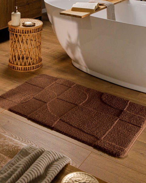 Rechteckiger brauner Badeteppich mit geometrischem Muster auf Holzboden neben freistehender Badewanne und Korb-Beistelltisch.