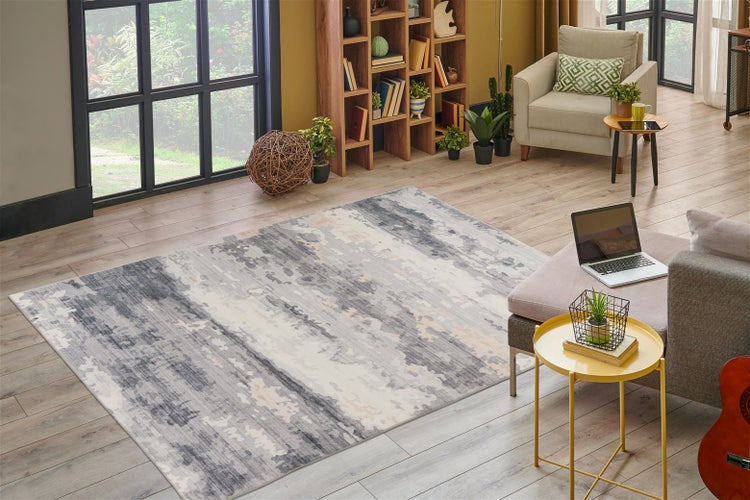 Rechteckiger Teppich mit abstraktem Muster in Grau und Creme in einem hellen Wohnzimmer mit Holzboden, Bücherregal und modernem Sessel.