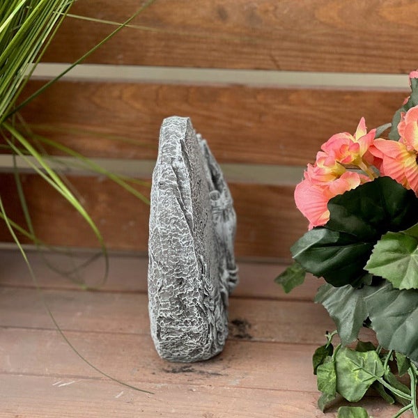 Steinfigur in Gedenksteinform mit Relief auf einem Holzuntergrund neben Blumen