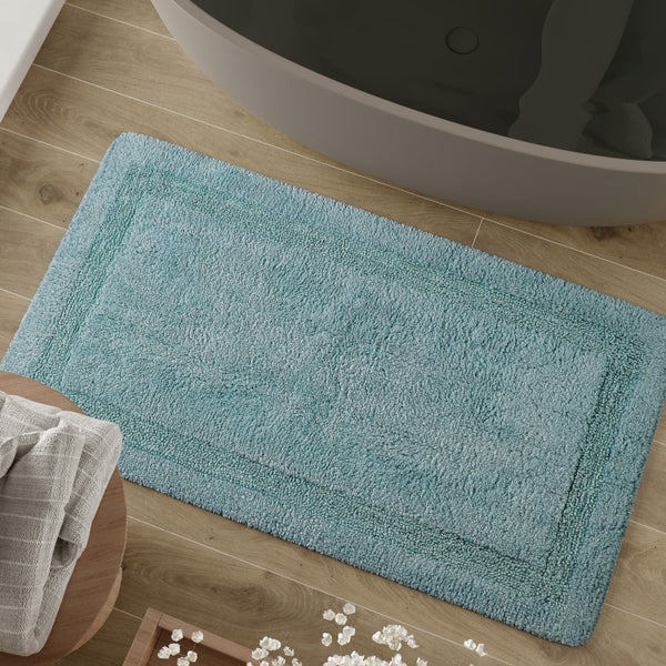 Türkisfarbene Badematte mit Bordüre auf Boden in Holzoptik neben einer Badewanne.