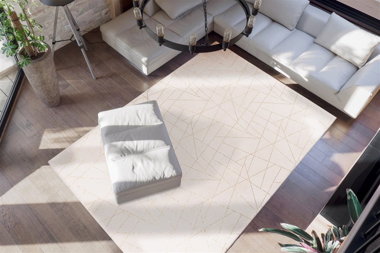Rechteckiger Teppich mit geometrischem Linienmuster in Gold auf hellem Grund in einem modernen Wohnzimmer mit grauem Sofa und Holzboden.