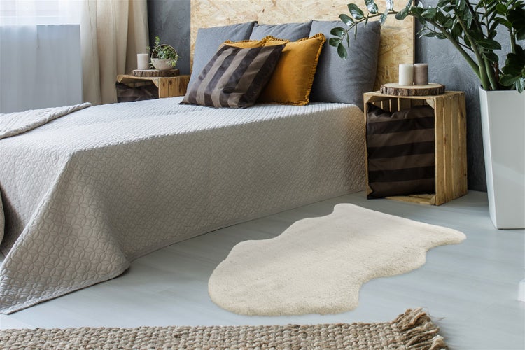 Weißes Kunstfell in einem hellen Schlafzimmer vor einem Bett mit grauer Tagesdecke. Rustikale Einrichtung mit Holzkisten und Pflanzen.