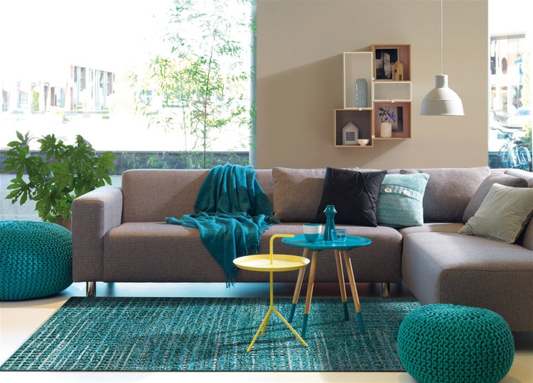 Modernes Wohnzimmer mit grauem Ecksofa, türkisfarbenem Teppich, Strickpoufs und bunten Beistelltischen vor einem hellen Fenster.