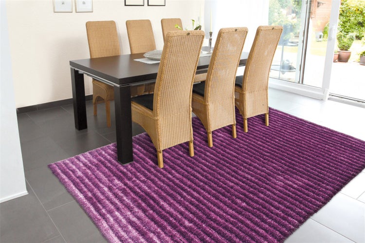 Rechteckiger Hochflor-Teppich mit Streifenmuster in Violetttönen unter einem dunklen Esstisch mit Korbstühlen in einem hellen Raum.