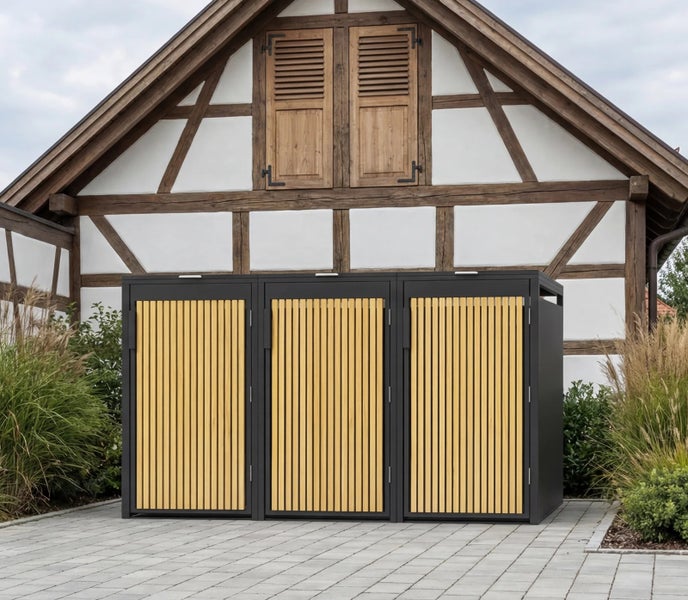 Dreifach-Mülltonnenbox aus Metall mit Holzlamellen vor einem Fachwerkhaus auf einer gepflasterten Fläche.