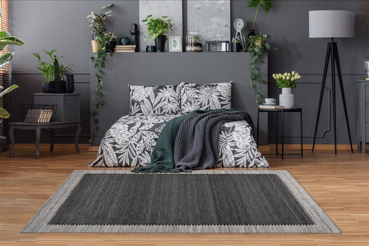 Moderner Teppich mit dunkelgrauem Zentrum und hellem Rand vor einem Bett mit Blattmuster-Bettwäsche und vielen Zimmerpflanzen.