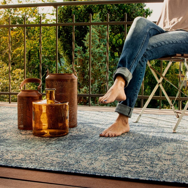 Outdoor-Teppich mit blau-weißem Vintage-Muster auf einem Holzbalkon mit Laternen, einem Glasgefäß und einer sitzenden Person.