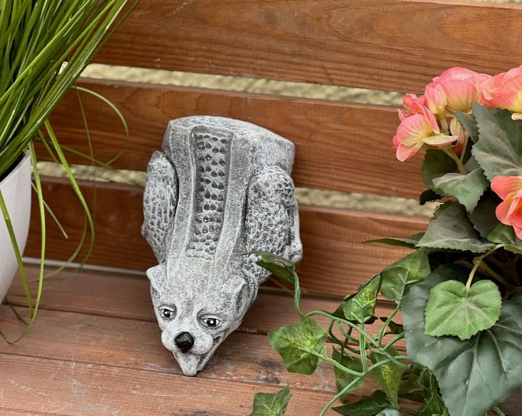 Steinfigur Gargoyle zum Einhängen an Holzzaun, dekorative Gartenfigur zwischen Pflanzen.
