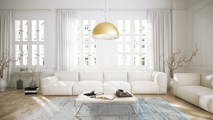 Helles, geräumiges Wohnzimmer mit weißem Ecksofa, goldener Pendelleuchte, Holztisch auf blauem Teppich und großen Fenstern mit Vorhängen.