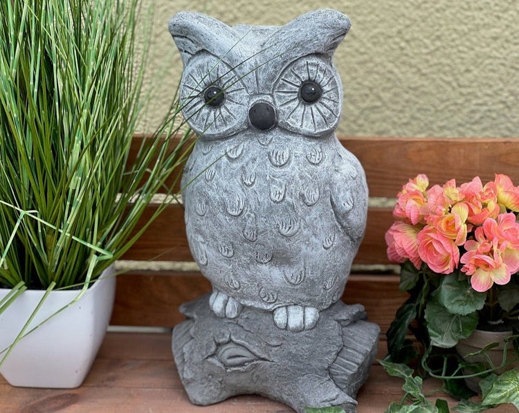 Dekorative Eulen-Gartenfigur in Steinoptik auf einem Ast, platziert zwischen grünem Ziergras und rosa Blumen auf einer Holzbank.