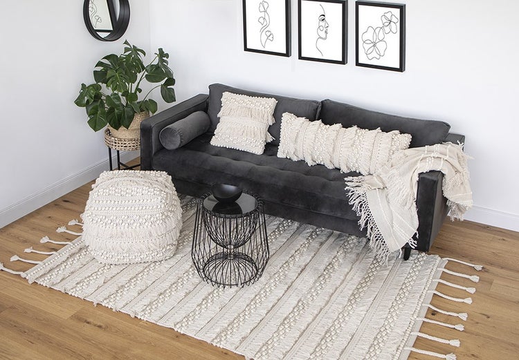 Modernes Wohnzimmer mit dunkelgrauem Sofa, cremefarbenen Makramee-Kissen, passendem Pouf und strukturiertem Teppich auf Holzboden.