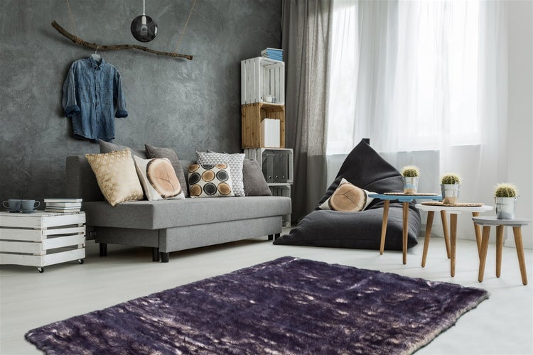 Modernes Wohnzimmer mit grauem Sofa, lila Kunstfellteppich, dunkel strukturierter Wand und verschiedenen Holzmöbeln.