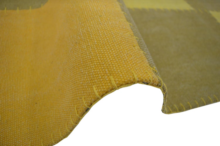 Nahaufnahme eines gelben Teppichs im Patchwork-Stil mit grober Webstruktur und sichtbarer Randnaht.