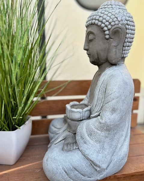Sitzende Buddha-Statue aus grauem Stein mit kleiner Schale, neben einer grünen Pflanze auf einer Holzbank.
