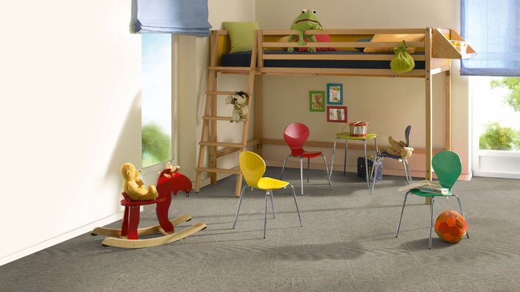 Helles Kinderzimmer mit Hochbett aus Holz, bunten Stühlen und einem Schaukelpferd auf grauen, strukturierten Teppichfliesen.