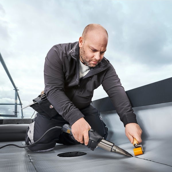 Ein Handwerker verschweißt eine Dachbahn mit einem Heißluftföhn und einer Andruckrolle auf einem Flachdach.