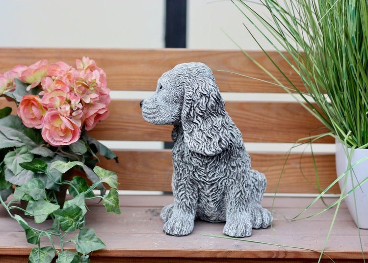 Gartenfigur in Form eines sitzenden Cockerspaniels auf einer Holzbank, neben einer Pflanze mit Blüten und einem Topf mit Gras.