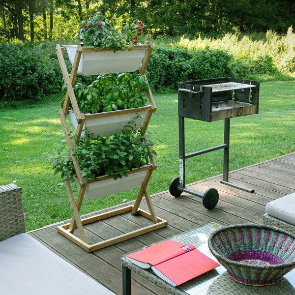 Vertikales Etagenbeet aus Holz mit drei weißen Pflanzkästen auf einer Holzterrasse neben einem Holzkohlegrill und Lounge-Möbeln.