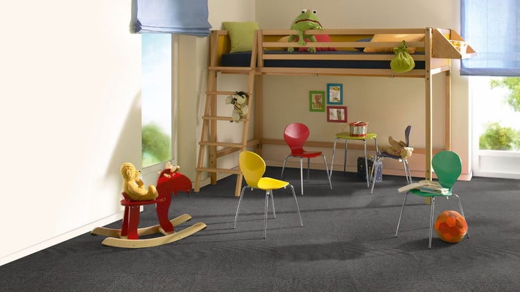 Ein helles Kinderzimmer mit dunklen Teppichfliesen, einem Hochbett aus Holz, farbenfrohen Stühlen und einem roten Schaukelpferd.