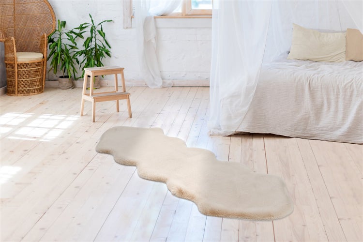 Langer beiger Kunstfellteppich mit welligen Kanten auf hellem Holzboden in einem hellen Zimmer mit Bett und Korbstuhl.