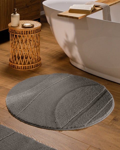 Runder grauer Badeteppich mit Wellenstruktur auf Holzboden neben einer Badewanne und einem Rattantisch.
