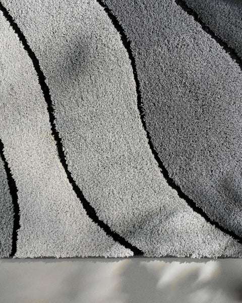 Hochflor-Teppich mit Wellenmuster in verschiedenen Grautönen und schwarzen Trennlinien.