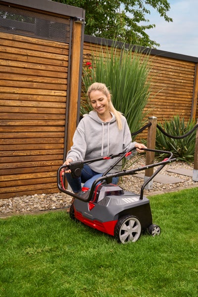 Eine Frau kniet auf einem Rasen im Garten und hält den Griff eines AL-KO Akku-Vertikutierers und Lüfters.