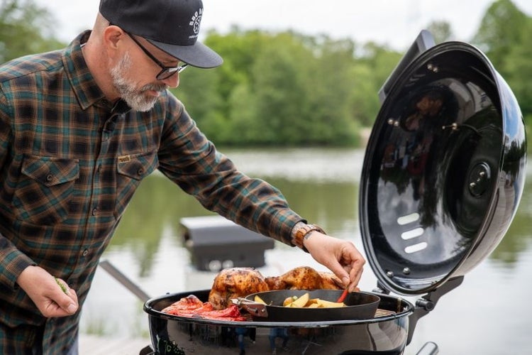 Ein Mann grillt ganze Hähnchen und Gemüse auf einem Kugelgrill im Freien an einem See.