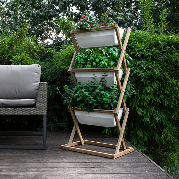Vertikales Pflanzregal aus Holz mit vier weißen Wannen, bepflanzt mit Kräutern und Tomaten, auf einer Holzterrasse neben einem Gartensessel.