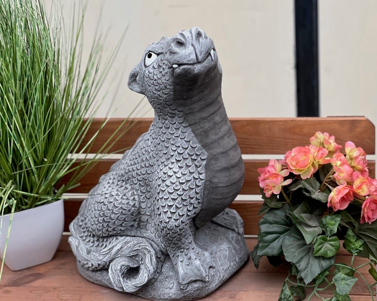 Gartenfigur Drache aus Steinguss blickt nach oben, platziert auf einer Holzbank zwischen einer Topfpflanze und Blumen.