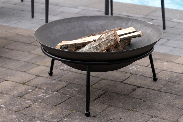 Runde Metall-Feuerschale auf schwarzem Dreibeingestell mit Brennholz auf einer gepflasterten Terrasse.