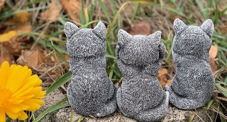 Drei Katzenfiguren als Gartendekoration in einer Reihe, sitzen auf einem Stein im Garten neben einer gelben Blume.