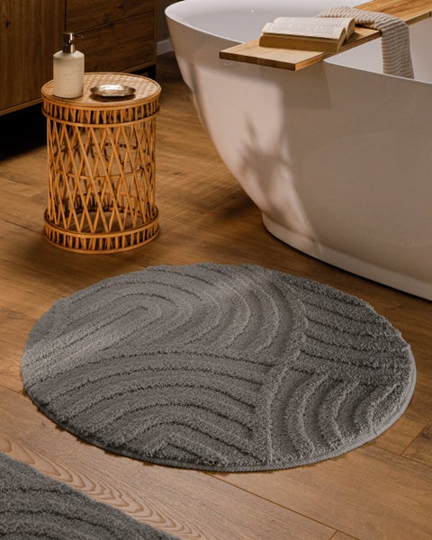 Runder grauer Badevorleger mit Wellenstruktur auf Holzboden neben einer weißen Badewanne und einem geflochtenen Beistelltisch.