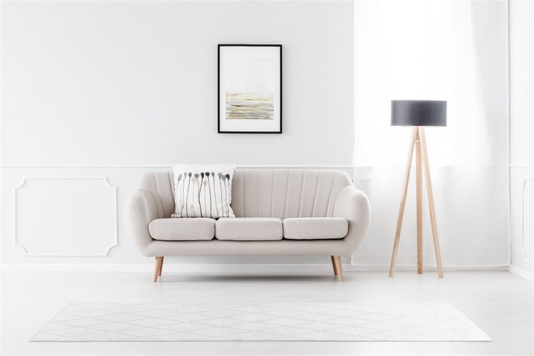 Helles Wohnzimmer mit beigem Sofa, Stativ-Stehlampe aus Holz, Wandbild und gemustertem Teppich vor einer weißen Wand mit Zierleisten.