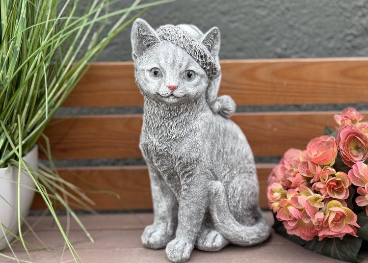 Dekorative Katzenfigur aus Stein für den Garten in sitzender Haltung mit kleiner Mütze auf dem Kopf.