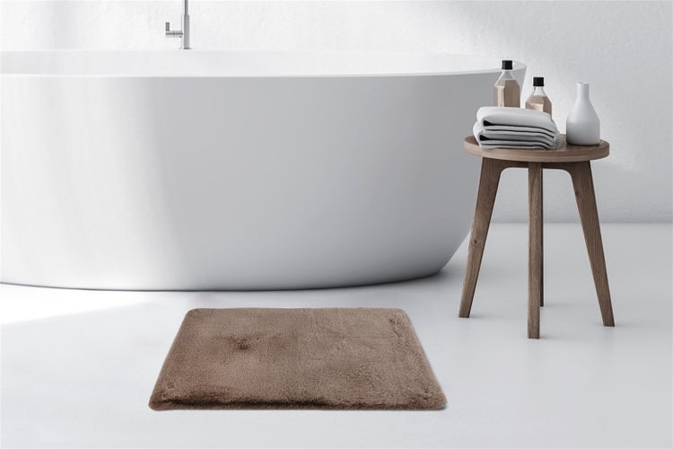 Flauschige braune Badematte vor einer freistehenden weißen Badewanne in einem hellen Badezimmer mit Holzschemel und Handtüchern.