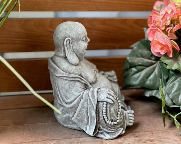 Sitzende Buddha-Figur in Stein-Optik als Gartendekoration auf einem Holztisch neben Blumen.