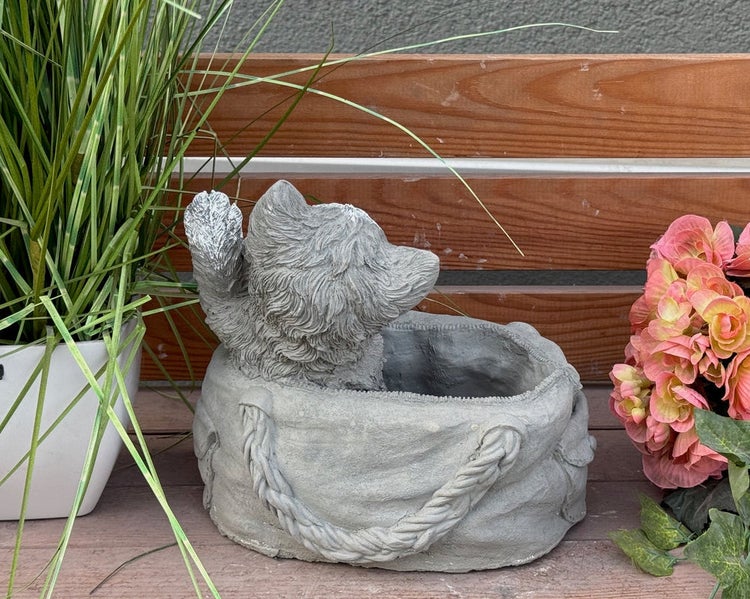 Dekorativer Pflanztopf aus grauem Steinguss in Form einer Tasche mit herausschauender Hundefigur auf einer Holzbank.