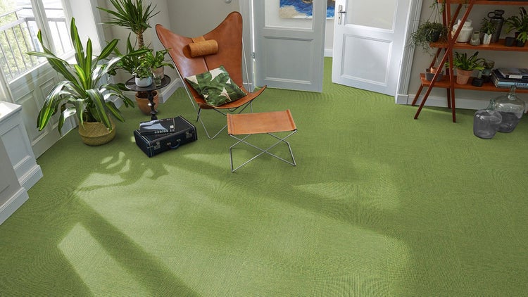 Modernes Wohnzimmer mit grünen Teppichfliesen, ledernem Butterfly-Sessel und Zimmerpflanzen in hellem Ambiente.