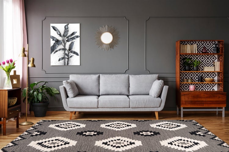 Modernes Wohnzimmer mit grauem Sofa, geometrischem Teppich, Holzregal und dunkelgrauer Wand mit Zierleisten und botanischem Bild.