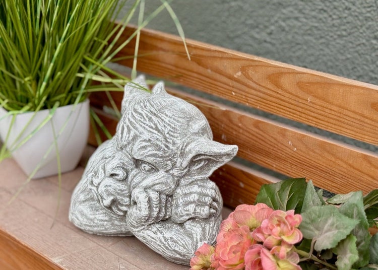 Dekorative Gartenfigur in Form eines Wasserspeiers aus Stein auf einer Holzoberfläche mit Pflanzen platziert.