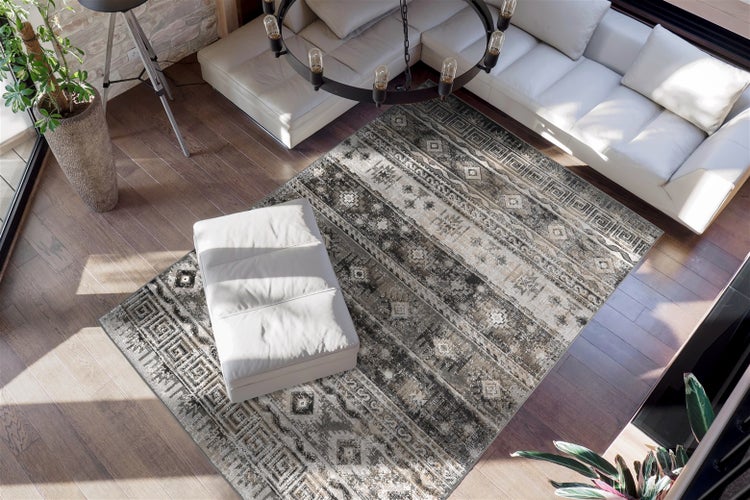 Hell gestalteter Wohnraum mit rechteckigem Webteppich mit Ethno-Muster auf dunklem Parkett, weißem Ecksofa, Hocker und großem Kronleuchter.
