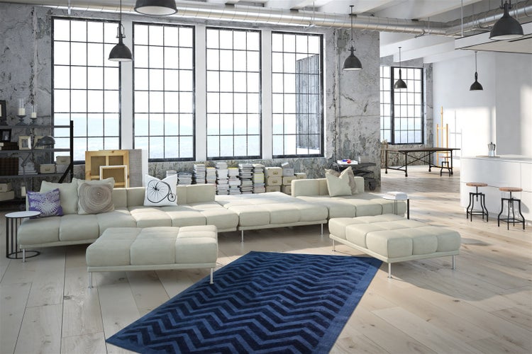 Wohnzimmer im Industriestil mit beigem Modulsofa, dunkelblauem Teppich mit Zickzack-Muster auf hellem Holzboden und großen Fenstern.