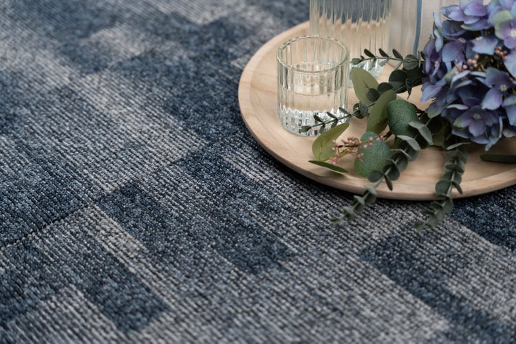 Gemusterter Teppich in Blau und Grau mit gerippter Struktur, dekoriert mit einem Holztablett, Glasgefäß und Blumen.