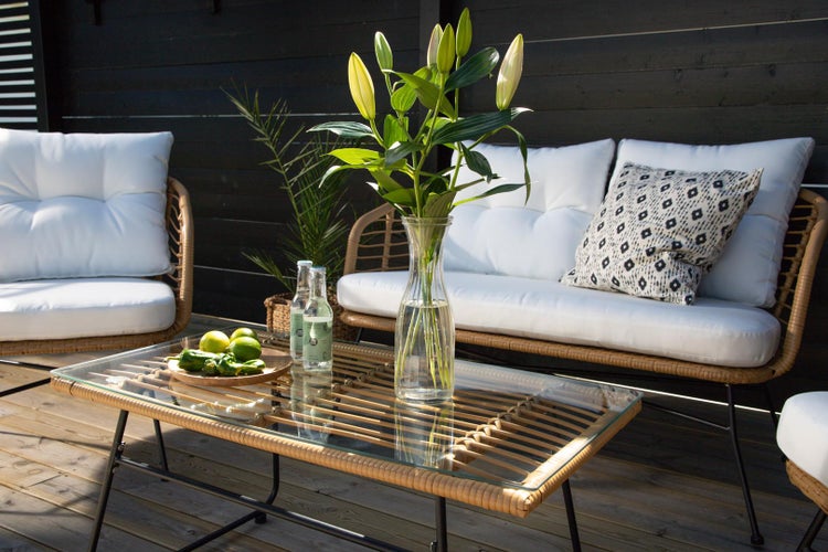 Terrassenszene mit Rattansitzgruppe, bestehend aus Sofa, Sessel und Tisch mit Glasplatte, dekoriert mit Blumen in Vase, Getränken und Früchten.