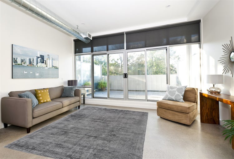 Helles, modernes Wohnzimmer mit grauem Sofa, grauem Teppich, braunem Sessel und großen Glasschiebetüren, die auf einen Balkon führen.